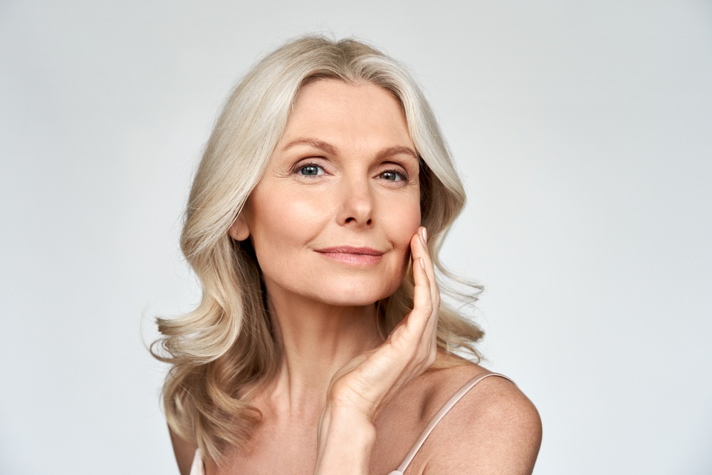Portrait of a mature woman with long, wavy silver hair gently touching her face, showcasing a natural and radiant complexion against a soft, neutral background. Ideal for beauty and skincare topics.