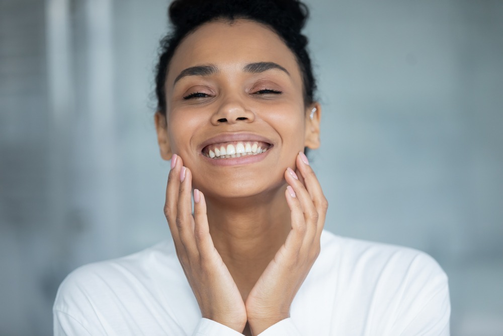 Smiling woman with curly hair in a white shirt gently touching her face, showcasing healthy skin and a joyful expression. Ideal for beauty and skincare topics.