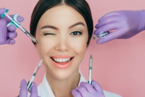 A woman with brown hair winks and smiles while four filler needles held by doctors’ hands in purple gloves surround her face.