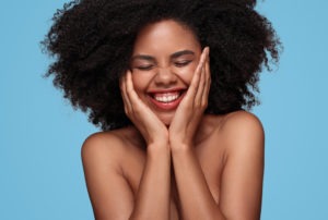 A confident young black woman holds her face and smiles with her eyes closed.
