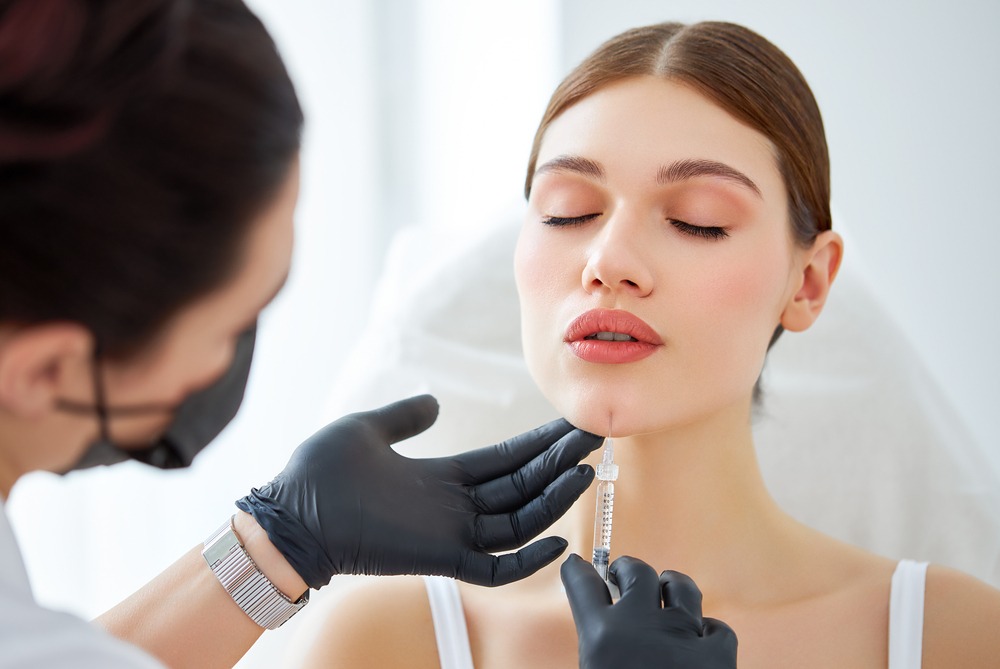 A medical professional in black gloves administers a cosmetic injection to a woman's chin in a bright, clinical setting. The woman, with her eyes closed, appears relaxed and is receiving a beauty treatment focused on enhancing facial features.