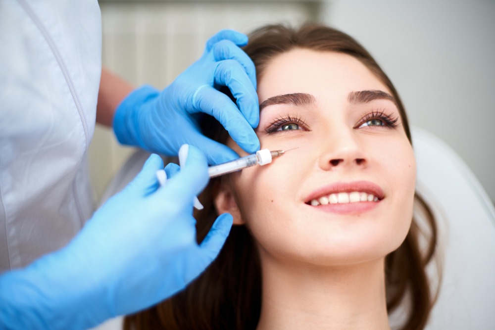 Aesthetic treatment session showing a dermatologist administering an injectable procedure to a smiling young woman in a clinical setting. The practitioner wears blue gloves and focuses on the eye area, highlighting popular cosmetic procedures like dermal fillers or Botox.