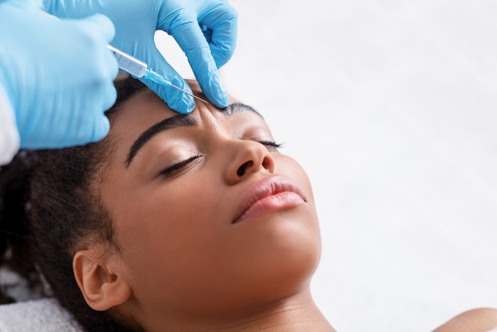 Close-up of a woman receiving a cosmetic injection on her forehead, performed by a professional wearing blue gloves, in a clinical setting focused on skincare treatments.