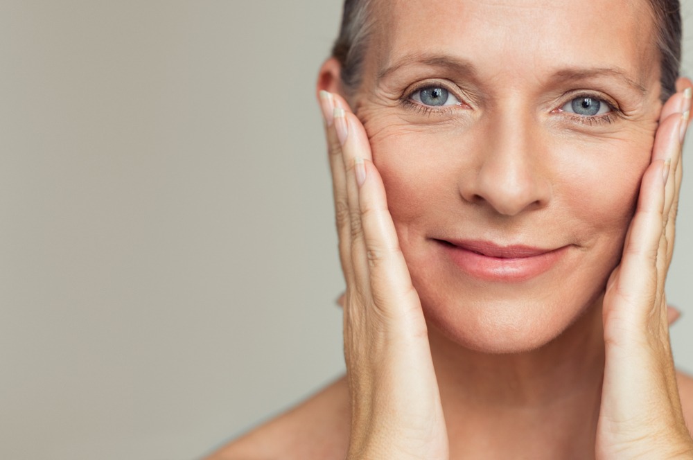 Smiling mature woman gently touching her face, showcasing healthy skin and a natural glow, against a neutral background. Ideal for beauty and skincare topics.