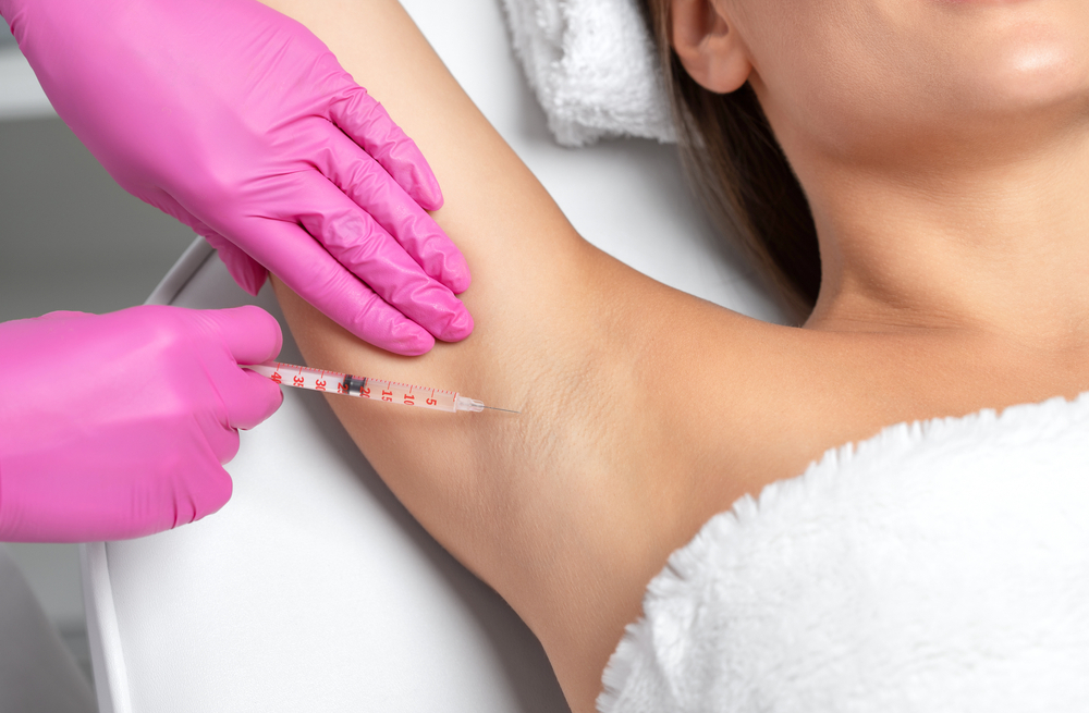Medical professional administering an underarm injection using a syringe, wearing pink gloves, in a clinical setting. The patient is relaxed on a treatment table, highlighting aesthetic procedures or medical treatments related to underarm care.