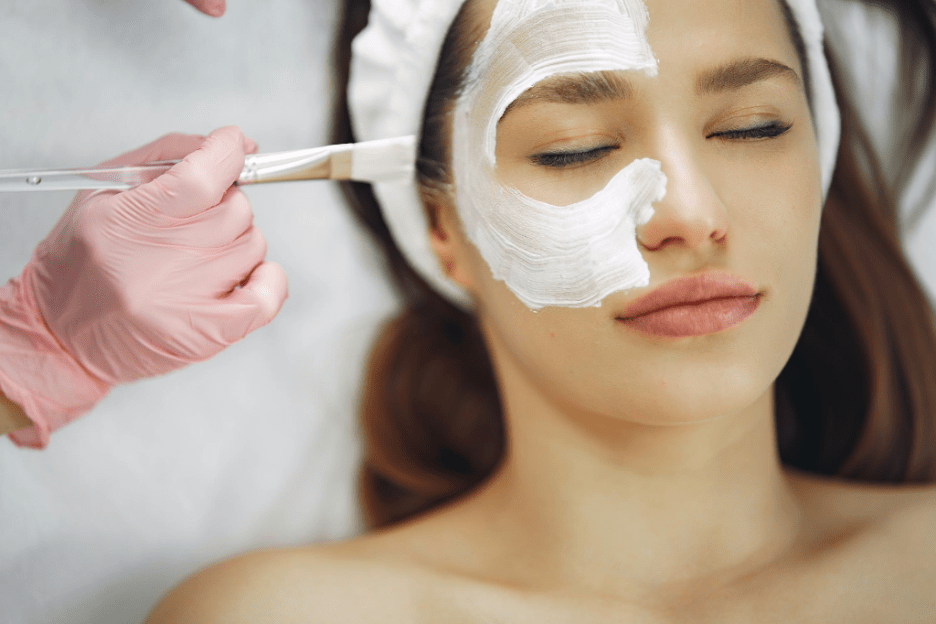 Woman receiving a facial treatment with a white mask applied to her face by a beautician wearing pink gloves, showcasing a relaxing spa environment.