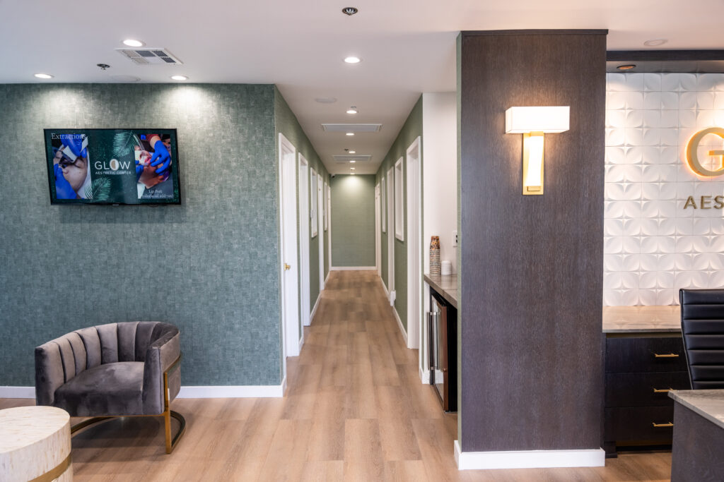 Modern aesthetic clinic interior featuring a welcoming reception area, green textured walls, and a hallway leading to treatment rooms. A flat-screen TV displays aesthetic treatment information, while stylish seating and elegant lighting enhance the ambiance.