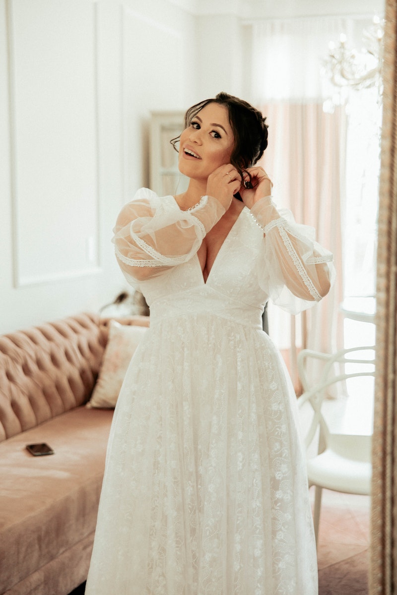 Bride getting ready in a beautiful white wedding dress with sheer puff sleeves, adjusting her earrings in a well-lit room with soft furnishings and a mirror.