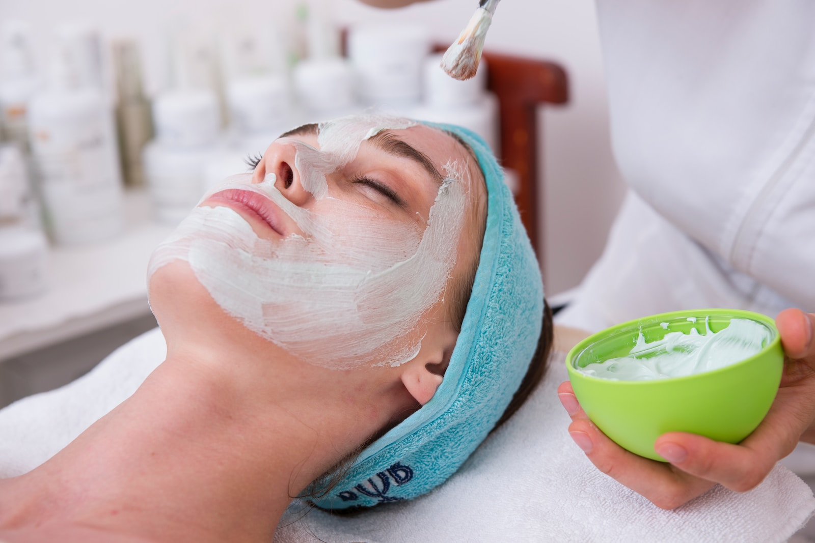 Woman receiving a facial treatment with a white mask applied to her face, while a beautician holds a green bowl of cream. The background features various skincare products on shelves, emphasizing a spa environment.