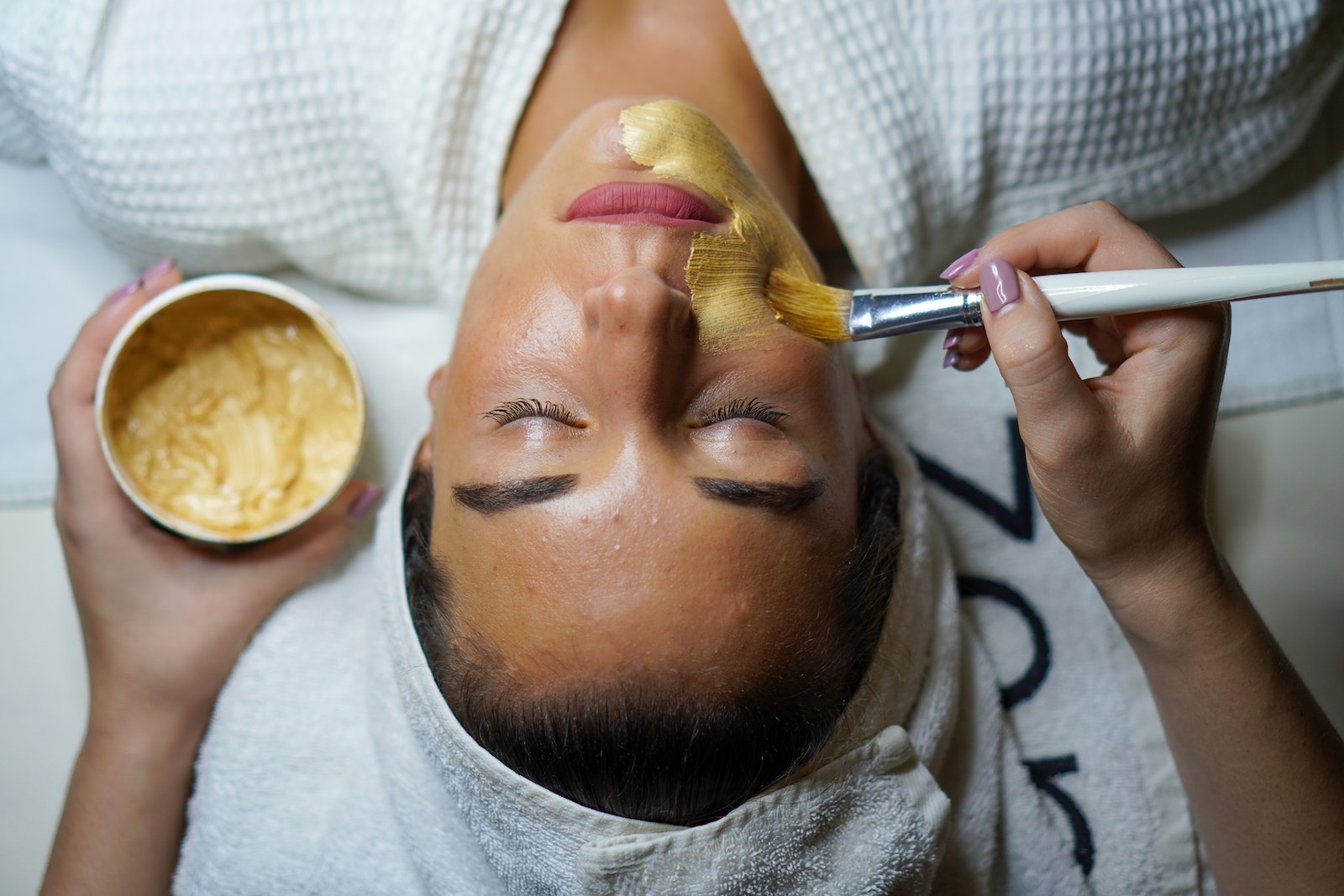 A woman receives a facial treatment as a beautician applies a golden mask using a brush, with a container of the mask visible in the background. The setting features a spa-like atmosphere, emphasizing skincare and relaxation.