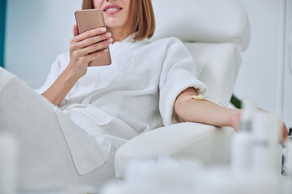 Woman in a white robe relaxing in a treatment chair, using a smartphone while receiving an IV infusion, with medical supplies in the background.