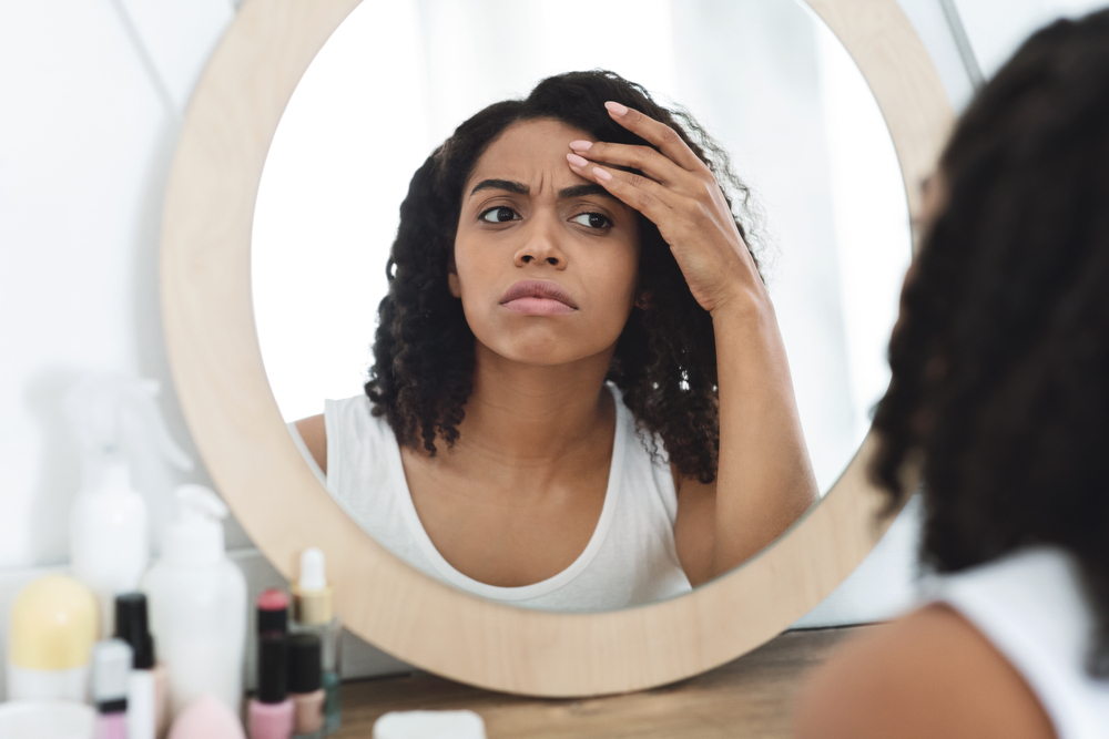 A woman with curly hair looks concerned while examining her reflection in a round mirror, with various skincare and cosmetic products visible on the table in front of her.
