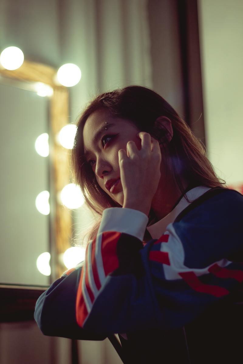 A young woman wearing a colorful athletic jacket listens to music with headphones, gazing thoughtfully while illuminated by soft, warm lighting from a nearby mirror.
